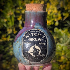 May include: A round ceramic bottle with a cork stopper, featuring a blue and purple mottled glaze. A black label on the front displays the text "WITCH'S BREW SINCE 1692 POTION NO 13" and a graphic of a witch flying on a broomstick against a full moon.