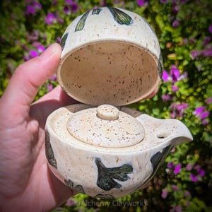 May include: A ceramic teapot with a speckled cream glaze and hand-painted dark green ginkgo leaf motifs. The teapot features a removable lid with a small knob and a side-mounted spout. The text Alchemy Clayworks is visible at the bottom.