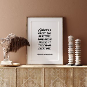 May include: A framed print with a white background and black text that reads "There's a great, big, beautiful tomorrow shining at the end of every day." The text is in a stylized font. The print is on a wooden shelf with a brown background. There are two stacks of white stones on the shelf.
