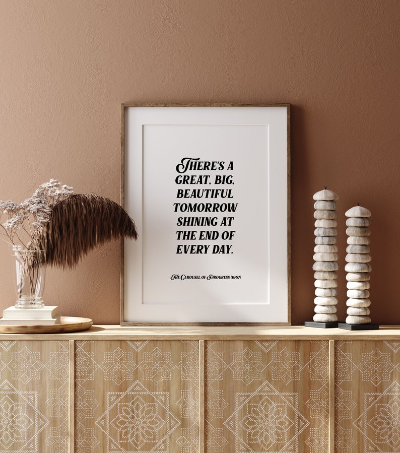 May include: A framed print with a white background and black text that reads "There's a great, big, beautiful tomorrow shining at the end of every day." The text is in a stylized font. The print is on a wooden shelf with a brown background. There are two stacks of white stones on the shelf.