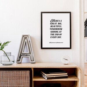 May include: A black framed print with a white background and black text that reads "There's a great, big, beautiful tomorrow shining at the end of every day." The print is hanging on a white wall above a wooden shelf with a gray concrete letter A, a clear glass vase with a green plant, and a white clock.