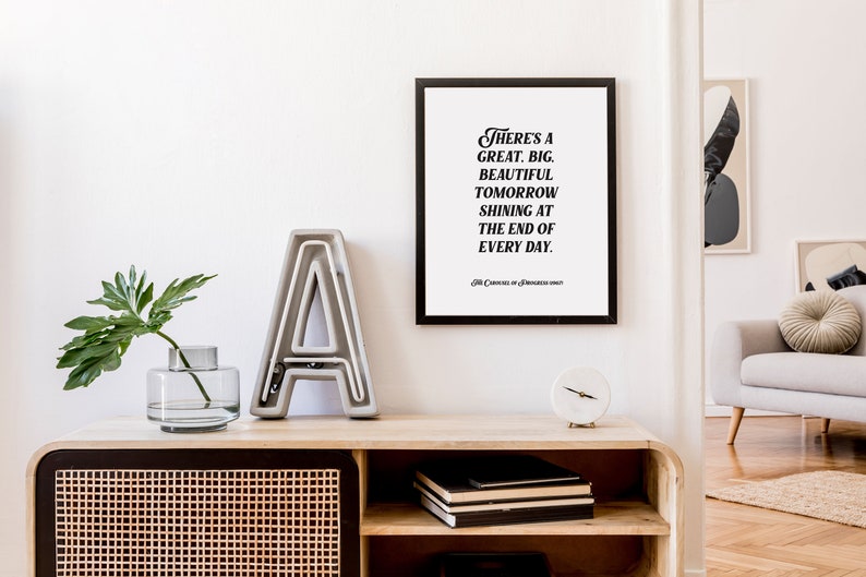 May include: A black framed print with a white background and black text that reads "There's a great, big, beautiful tomorrow shining at the end of every day." The print is hanging on a white wall above a wooden shelf with a gray concrete letter A, a clear glass vase with a green plant, and a white clock.