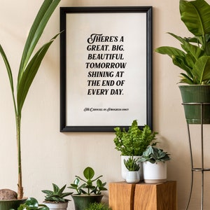 May include: A framed print with a black frame and a white background. The print features a quote in black text that reads "There's a great, big, beautiful tomorrow shining at the end of every day. The Carousel of Progress (1967)". The print is displayed on a wall with several potted plants in front of it.
