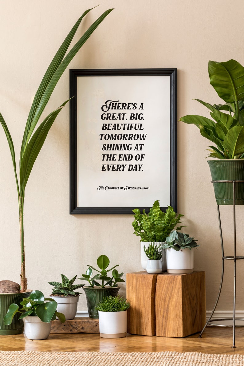 May include: A framed print with a black frame and a white background. The print features a quote in black text that reads "There's a great, big, beautiful tomorrow shining at the end of every day. The Carousel of Progress (1967)". The print is displayed on a wall with several potted plants in front of it.