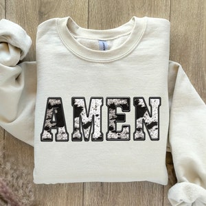May include: A beige sweatshirt with the word "AMEN" printed on the front in black and white. The letters are in a bold, blocky font.