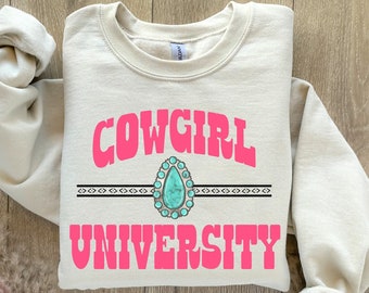 Könnte beinhalten: Ein beiger Pullover mit dem Text "COWGIRL UNIVERSITY" in rosa Schrift. Ein türkisfarbener Edelstein mit silbernen Akzenten ist mittig über dem Text platziert.
