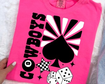 May include: Pink t-shirt with a black and white graphic design featuring the word "COWBOYS" in a retro font, a spade, a star, and dice.