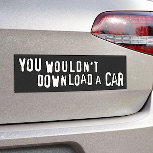 Peut inclure: Un aimant de voiture rectangulaire noir avec le texte blanc "YOU WOULDN'T DOWNLOAD A CAR" est fix&eacute; &agrave; l'arri&egrave;re d'une voiture argent&eacute;e. L'aimant est positionn&eacute; sous le feu arri&egrave;re.