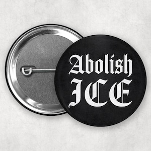 Puede incluir: Botón negro con el texto "Abolish ICE" en letras góticas blancas. El botón es redondo y tiene una parte trasera de metal plateado con un alfiler. Declaración política.