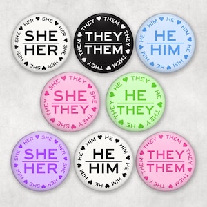 Könnte beinhalten: Eine Sammlung runder Anstecknadeln mit Pronomen in verschiedenen Farben, darunter Weiß, Schwarz, Blau, Rosa, Grün und Lila. Jede Nadel zeigt Pronomen wie "She/Her", "He/Him" und "They/Them" in einer klaren Schrift, umgeben von Herzen.