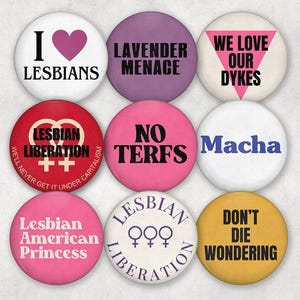 May include: Nine circular badges with slogans and symbols. The badges display text such as "I LESBIANS", "LAVENDER MENACE", and "WE LOVE OUR DYKES". The colours are white, purple, red, pink, blue, and yellow.