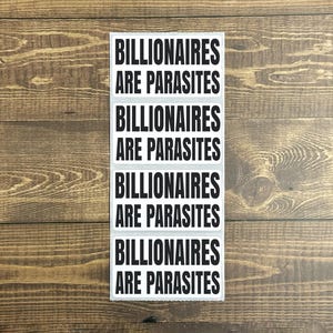 Könnte beinhalten: Ein weißer, rechteckiger Aufkleber mit dem Text "BILLIONAIRES ARE PARASITES" in fetten, schwarzen Großbuchstaben, fünfmal wiederholt. Der Aufkleber befindet sich vor einem Holzgrund.