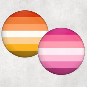 以下が含まれることがあります： プライドフラッグのデザインが施された2つの丸いボタン。1つのボタンは、オレンジ、黄色、白のストライプのグラデーションが特徴です。もう1つのボタンは、ピンクと白のストライプです。どちらのボタンも光沢のある仕上がりです。