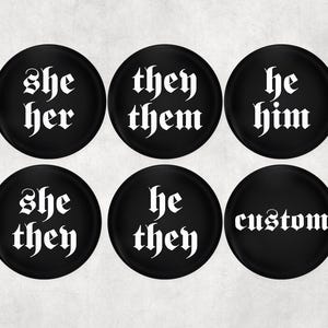 May include: Six black round buttons with white gothic font text. The buttons display the pronouns "she/her", "they/them", "he/him", "she/they", "he/they", and "custom". These buttons are a statement of identity.