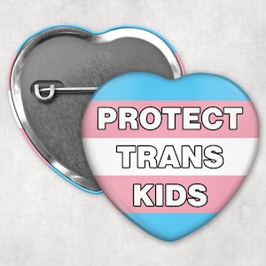 May include: Heart-shaped button with the text "PROTECT TRANS KIDS" in black, outlined in white, over a light blue, white, and pink background. The button has a pin back and a silver metal backing. The design is a symbol of support.