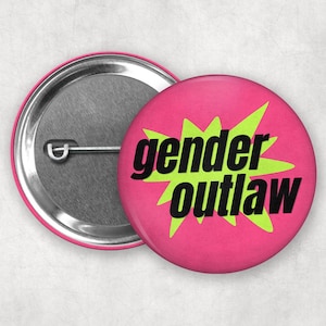 May include: A pink button with the words "gender outlaw" in black text over a green starburst design. The button has a silver metal back with a pin.