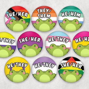 Puede incluir: Un conjunto de botones redondos y coloridos con pronombres y un diseño de rana de dibujos animados. Cada botón muestra un conjunto de pronombres diferente, como "Ella/Ella", "Ellos/Ellas" y "Él/Él", con banderas del orgullo.