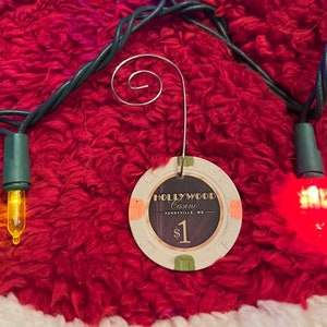 Könnte beinhalten: Ein Weihnachtsornament in Form eines Casino-Chips mit dem Text "Hollywood Casino Perryville, MD" und "$1". Der Ornament hängt an einem roten Weihnachtsbaumrock mit weißem Besatz und ist von Lichterketten umgeben.