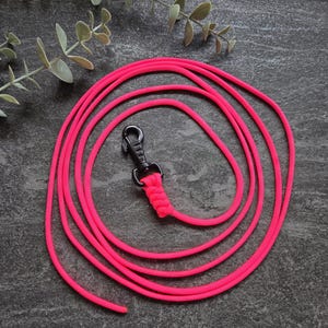 Op de afbeelding: Een felroze hondenriem gemaakt van gevlochten koord. De riem heeft een zwarte metalen clip en een gevlochten lus. De riem is opgerold op een donkergrijze ondergrond.