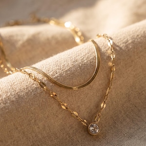 May include: A layered gold-tone necklace featuring two distinct chains. The top chain is a flat, flexible herringbone style, while the bottom chain consists of oval-shaped links and a single round, clear, bezel-set stone pendant.