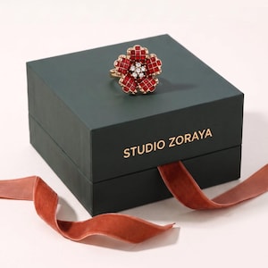 May include: A gold-toned ring shaped like a five-petaled flower, featuring red square-cut stones on the petals and a cluster of clear round stones in the center, resting on a dark green square box labeled STUDIO ZORAYA with a velvet ribbon.