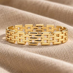 May include: A gold-toned metal bracelet featuring a repeating pattern of horizontal, rectangular links connected by smaller vertical bars. The bracelet has a polished, reflective surface and a structured, interlocking design that forms a continuous band.