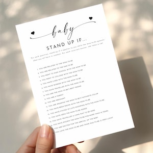 May include: A white rectangular card featuring black text titled Baby Stand Up If. The card lists eighteen numbered prompts for a party game, with two small black hearts flanking the word baby at the top. The text is printed in a clear, sans-serif font.
