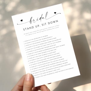 May include: A white card with the words "bridal STAND UP, SIT DOWN" in black script. The card has a list of questions for a bridal shower game. The card is held by a person.