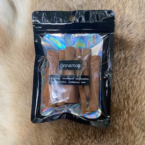 May include: A black, sealed package containing several cinnamon sticks. The package has a holographic interior and a label that reads "cinnamon" with additional text about its properties. The cinnamon sticks are a warm brown color.