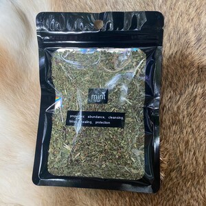 May include: A black, resealable bag filled with dried mint leaves. The bag has a clear window, displaying the green herbs. The word "mint" is printed on the bag, along with the properties: abundance, cleansing, focus, healing, and protection.