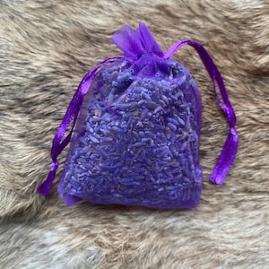 Op de afbeelding: Een kleine, paarse mesh zak gevuld met gedroogde lavendelbloemen. De zak is aan de bovenkant vastgebonden met een bijpassend paars lint. De lavendelbloemen zijn dieppaars van kleur en de zak ligt op een bruin, getextureerd oppervlak.