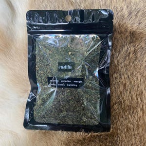 May include: A black, resealable bag filled with dried nettle leaves. The bag has a clear window, displaying the green herbs. Text on the bag reads "nettle" and lists properties: protection, strength, healing, justice, banishing.