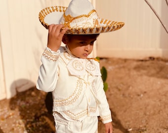 May include: A young child wearing a white charro suit with gold embroidery. The suit includes a vest, pants, and a wide-brimmed hat with gold trim.