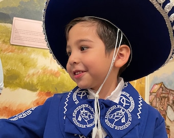 Puede incluir: Un niño pequeño vistiendo un traje de charro azul con bordados blancos. Lleva un sombrero azul con ribete plateado. El traje tiene una camisa blanca y una corbata de moño blanca. El niño sonríe.