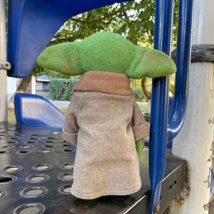 Puede incluir: Un juguete de peluche verde y marr&oacute;n que se asemeja a un personaje de la franquicia Star Wars, de pie sobre una superficie de metal negro con una estructura de metal azul detr&aacute;s.
