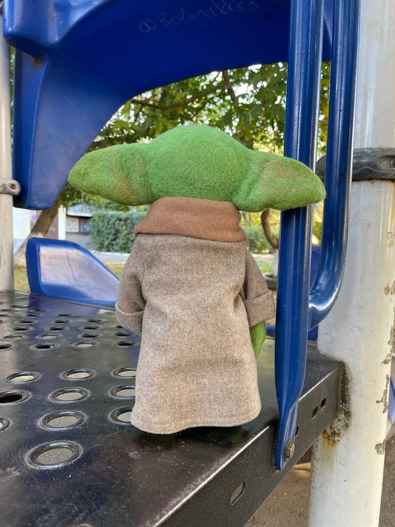 Puede incluir: Un juguete de peluche verde y marr&oacute;n que se asemeja a un personaje de la franquicia Star Wars, de pie sobre una superficie de metal negro con una estructura de metal azul detr&aacute;s.