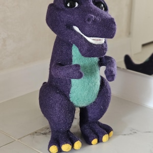 Op de afbeelding: Een paarse pluche dinosaurus met een groene buik en gele voeten. Het speelgoed heeft een vriendelijke glimlach en zwarte ogen. De dinosaurus staat op een betegeld oppervlak. Het speelgoed is ongeveer 30 cm hoog.