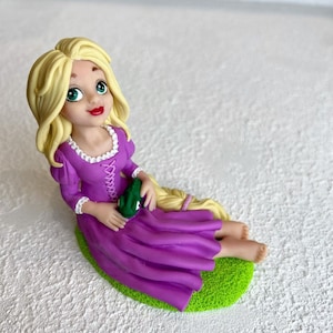 May include: A handmade figurine of Rapunzel from the Disney movie Tangled. She is wearing a purple dress with a white lace trim and holding a green frog. She has long blonde hair and is sitting on a green base.