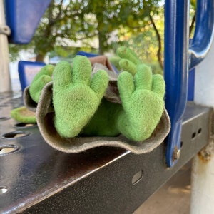 Puede incluir: Un t&iacute;tere de fieltro verde con manos grandes, acostado sobre una superficie de metal negro. El t&iacute;tere est&aacute; parcialmente oculto debajo de una tela marr&oacute;n.