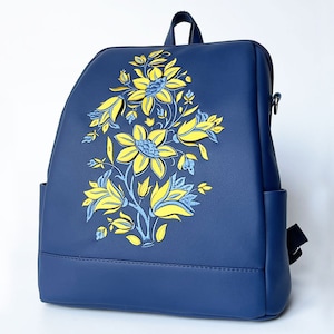 Puede incluir: Una mochila azul con un diseño floral amarillo y azul. La mochila tiene cierre de cremallera y correas ajustables.