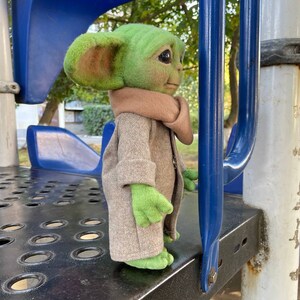 Puede incluir: Un juguete de peluche verde y marr&oacute;n que se asemeja a un personaje de la franquicia Star Wars, de pie en una estructura de juego de metal. El juguete lleva un abrigo marr&oacute;n y tiene orejas grandes.