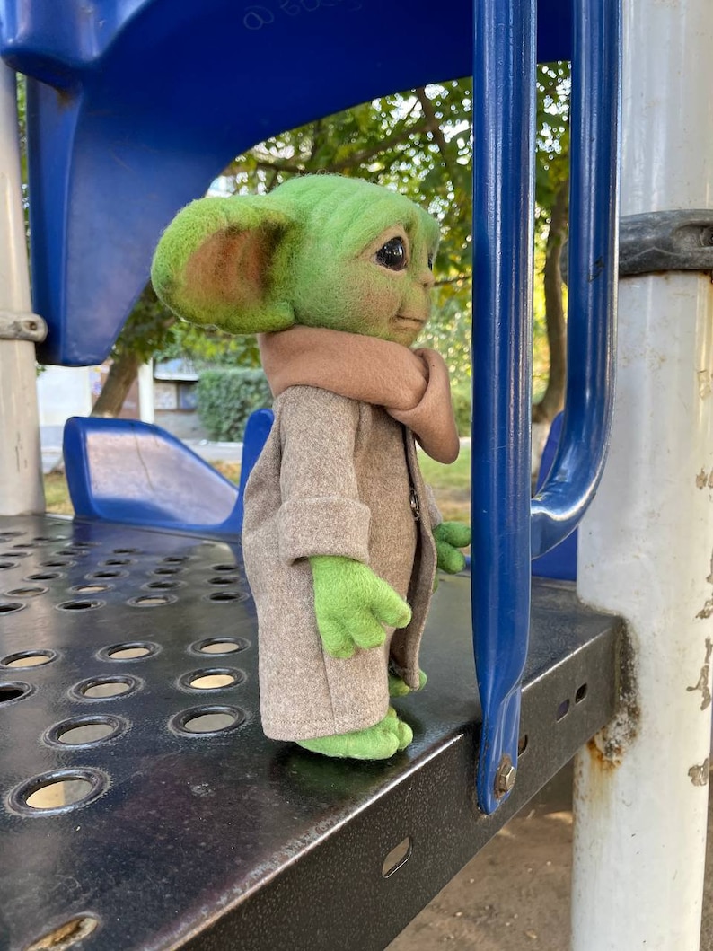 Puede incluir: Un juguete de peluche verde y marr&oacute;n que se asemeja a un personaje de la franquicia Star Wars, de pie en una estructura de juego de metal. El juguete lleva un abrigo marr&oacute;n y tiene orejas grandes.