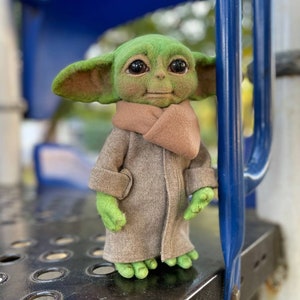 Puede incluir: Un juguete de fieltro verde de un personaje de la franquicia Star Wars, que lleva un abrigo de fieltro marr&oacute;n y una bufanda de fieltro marr&oacute;n. El juguete est&aacute; de pie sobre una superficie de metal negro.
