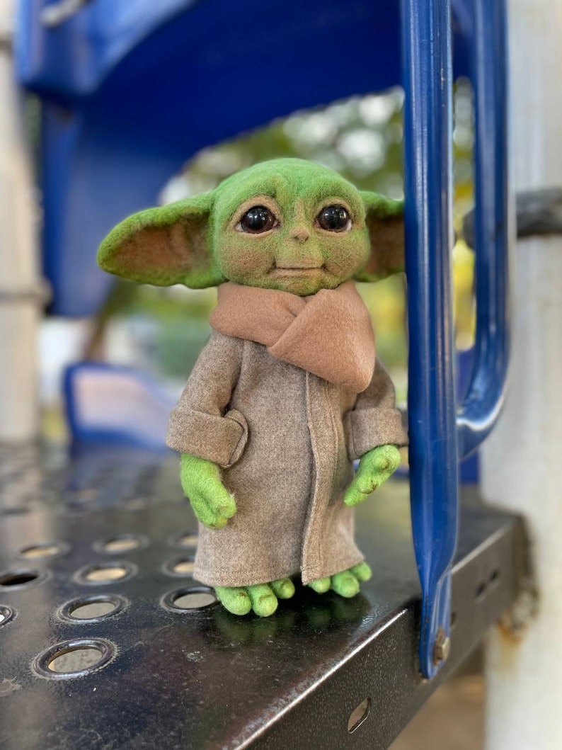 Puede incluir: Un juguete de fieltro verde de un personaje de la franquicia Star Wars, que lleva un abrigo de fieltro marr&oacute;n y una bufanda de fieltro marr&oacute;n. El juguete est&aacute; de pie sobre una superficie de metal negro.