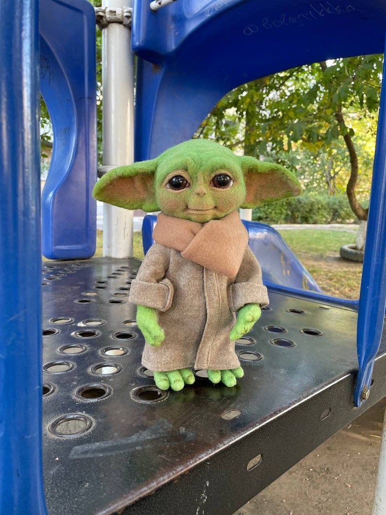 Puede incluir: Un juguete de peluche verde con ojos grandes y un abrigo marr&oacute;n, de pie sobre una superficie de metal con agujeros. El juguete lleva una bufanda marr&oacute;n y tiene una expresi&oacute;n amigable.