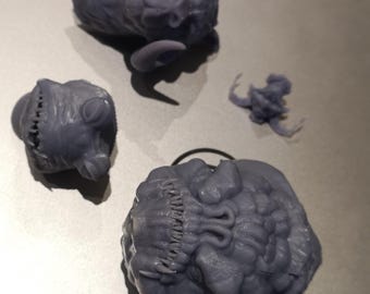 May include: Several small, gray, 3D-printed sculptures are arranged on a surface. The sculptures include a horned creature, a head with a stitched seam, and a small, abstract form. The details are intricate, with visible textures.