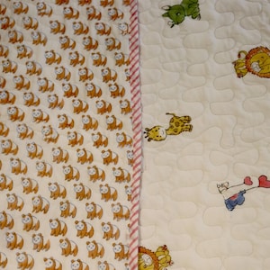 Pode incluir: Um edredom acolchoado para bebé com dois padrões distintos. Um lado apresenta um padrão repetido de pandas laranja e brancos. O outro lado tem um fundo branco com animais de desenho animado espalhados, incluindo um leão, uma girafa e um elefante.