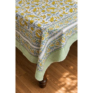 Può includere: Una tovaglia bianca con un motivo ripetuto di fiori gialli e viticci blu. Il tessuto presenta un'ampia bordura con un design a righe verticali verdi e bianche lungo il bordo inferiore, che ricade sull'angolo di un tavolo in legno.