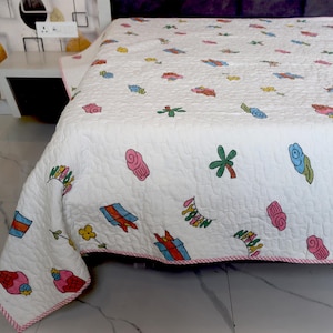 Op de afbeelding: Wit gewatteerd sprei met kleurrijke, grillige illustraties. Het ontwerp toont verschillende vormen en objecten in tinten roze, blauw, groen en oranje, met een roze rand. De sprei is gemaakt van een zacht, gewatteerd materiaal.
