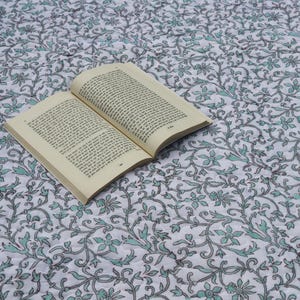 Può includere: Un libro aperto con pagine beige è appoggiato su una trapunta bianca con un motivo floreale in verde acqua e grigio. Un bordo di corda blu borda la trapunta. La scena suggerisce relax e lettura.